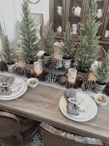Çanlar, kozalaklar, saksıdaki yılbaşı ağaçları ve ahşap ağaç süsleri ile gri peçetelerin yer aldığı modern rustik Noel masası.