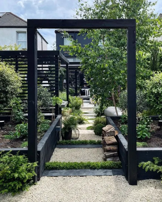 Siyah çerçeveli bahçe yatakları ve siyah pergola, bol yeşillik ve ağaçlarla birlikte serin ve modern İskandinav tarzı bir alan yaratır.