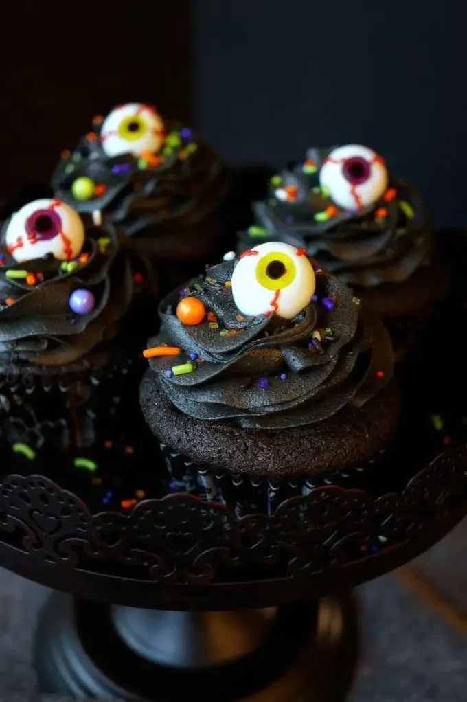 Üzerinde renkli konfetiler ve gözler bulunan siyah çikolatalı cupcakeler, Cadılar Bayramı partinizde harika misafir ikramları.
