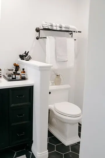 Siyah altıgen karolarla küçük bir çiftlik evi banyosu, tuvaleti ayıran yarım duvar ve siyah lavabo dolabı.