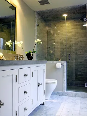 Beyaz duvarlar, yarım duvarlı gri karo duş alanı ve gri taş tezgahlı büyük beyaz lavabo ünitesiyle şık banyo.