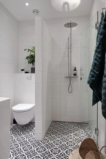 Beyaz uzun fayanslı duş alanı olan küçük banyo. Desenli fayans zemin, saksı bitkisi ve beyaz lavabo var.