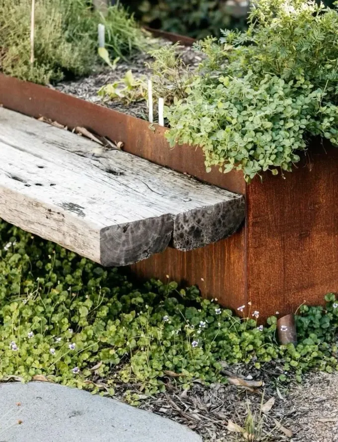 Bu metal saksı corten çelik kenarlıklara sahip; geri dönüştürülmüş ahşap bir ahşap bank da bulunuyor.