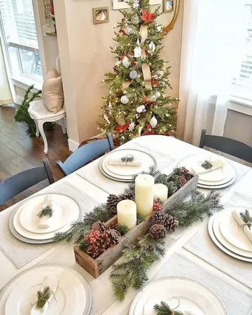 Gümüş tabak altlıkları, beyaz tabaklar, çam dalları, kozalaklar, meyveler ve mumların olduğu sade nötr bir Noel masa düzeni.