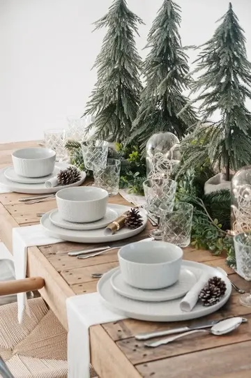 Mini ağaçlar, her zaman yeşil süsler, beyaz porselen, kozalaklar ve beyaz peçetelerle sıcak bir yılbaşı masası.