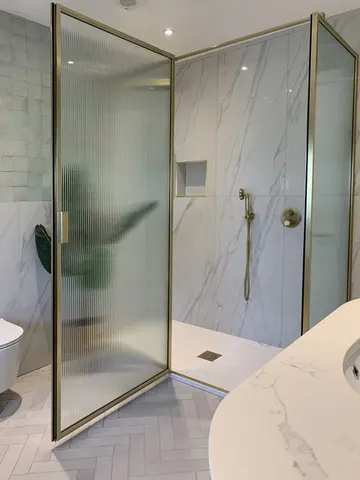 Beyaz mermer karolar, altın çerçeveli fitilli cam kapılar ve altın armatürlü şık banyo.