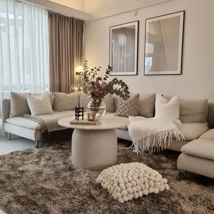 Nötr oturma odasında metal ayaklı U şeklinde köşe koltuk, nötr masa ve taupe tonlarında tekstiller bulunuyor.