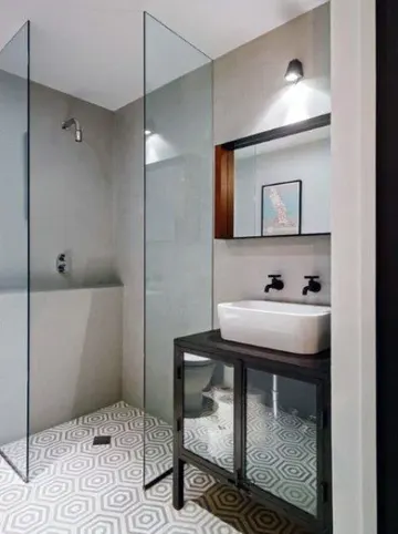 Beton duvarlı ve desenli zemin karolarıyla küçük bir banyo, küçük duş alanı, siyah lavabo ve tezgâh.